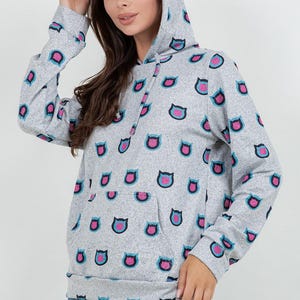 May include: A gray hooded sweatshirt with a repeating pattern of cat face shapes in pink, blue, and black. The sweatshirt has a front pocket and a drawstring hood. The model is wearing blue jeans.