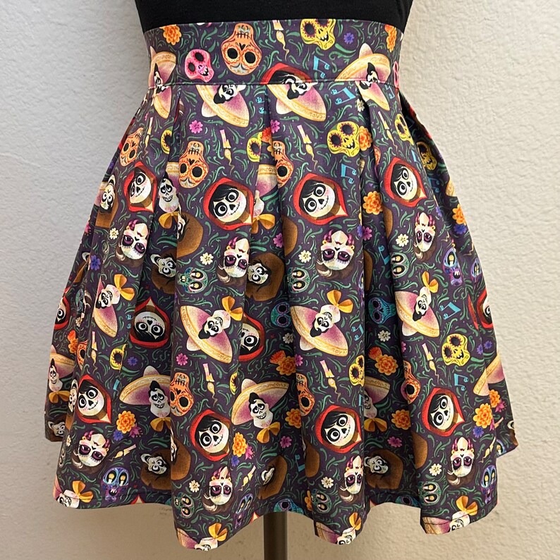 May include: A black pleated skirt with a colorful print of characters from the movie Coco. The print features skeletons, flowers, and musical notes on a dark background.