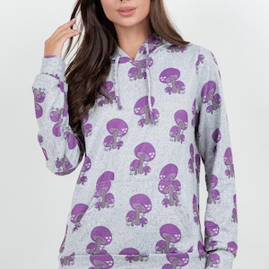 May include: A gray hoodie with a repeating pattern of purple mushrooms. The hoodie has a drawstring hood and a front pocket. The sleeves and hood trim are also gray with the same mushroom pattern. The model is wearing blue jeans.