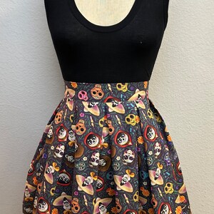 May include: A black sleeveless top with a pleated skirt featuring a colorful print of characters from the movie Coco. The skirt has a dark background with images of skeletons, flowers, and musical notes.