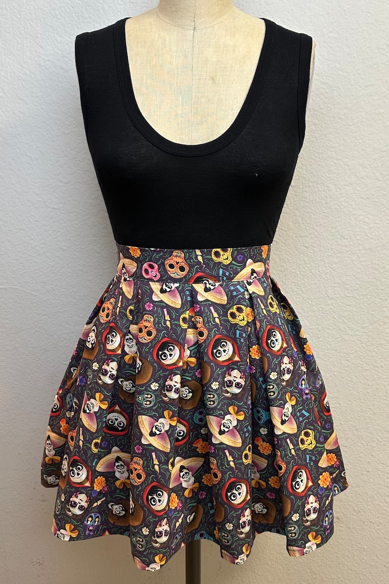 May include: A black sleeveless top with a pleated skirt featuring a colorful print of characters from the movie Coco. The skirt has a dark background with images of skeletons, flowers, and musical notes.