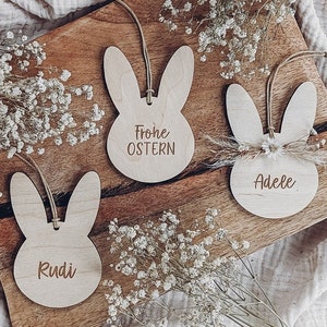 May include: Three wooden bunny-shaped Easter decorations with twine hangers. The top bunny says "Frohe Ostern" and the bottom two bunnies say "Rudi" and "Adele".