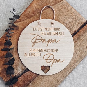 May include: A round wooden sign with the words "DU BIST NICHT NUR DER ALLERBESTE Papa SONDERN AUCH DER ALLERBESTE Opa" engraved in a stylish font. A heart shape is cut out at the bottom. A beige rope is attached for hanging.