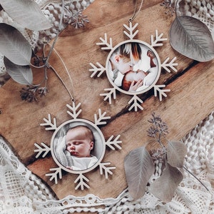 May include: Two wooden snowflake ornaments with photo inserts. One ornament features a photo of a baby and a sibling, while the other ornament features a photo of a baby.