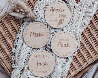 Puede incluir: Tres adornos redondos de madera con bordes de alfabeto. Cada adorno tiene un nombre y el texto "SCHULKIND" con una fecha. Los nombres son Paul, Noah y Thea.