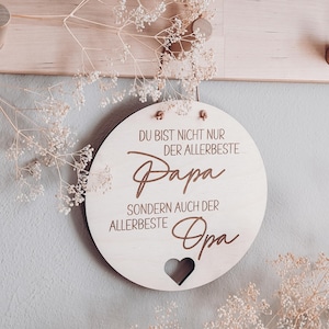 May include: A round wooden sign with the text "DU BIST NICHT NUR DER ALLERBESTE Papa SONDERN AUCH DER ALLERBESTE Opa" in brown script. A heart-shaped cutout is at the bottom. The sign is hanging on a light-colored wall.