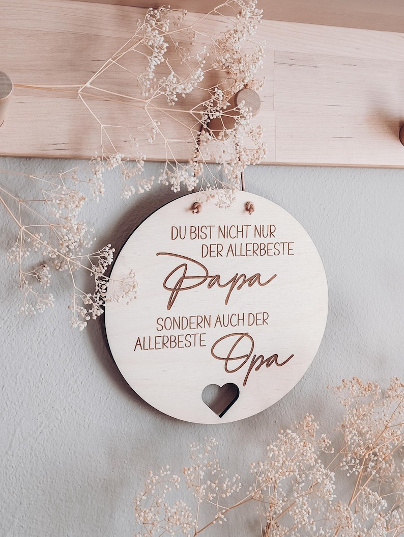May include: A round wooden sign with the text "DU BIST NICHT NUR DER ALLERBESTE Papa SONDERN AUCH DER ALLERBESTE Opa" in brown script. A heart-shaped cutout is at the bottom. The sign is hanging on a light-colored wall.