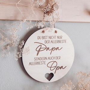 May include: A round wooden sign with the words "DU BIST NICHT NUR DER ALLERBESTE Papa SONDERN AUCH DER ALLERBESTE Opa" in brown script. A heart shape is cut out at the bottom. The sign is hanging on a light gray wall with dried flowers.