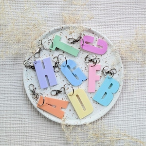 May include: A collection of pastel-colored acrylic letter keychains featuring the letters A, B, D, F, G, H, I, L, and T. Each letter is attached to a silver-toned metal split ring and a lobster claw clasp, arranged on a speckled white circular tray.
