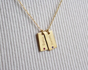 Op de afbeelding: Goudkleurige ketting met twee rechthoekige hangers, elk gegraveerd met een naam en een kleine edelsteen. De ketting wordt getoond tegen een witte, geribbelde stoffen achtergrond.