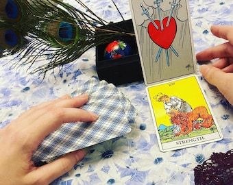 May include: Tarot cards spread out on a table with a peacock feather and a small decorative box. The cards show the Three of Swords and Strength.