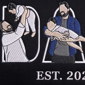 Puede incluir: Diseño bordado en negro con la palabra "DAD" e ilustraciones de un padre y un niño. El texto "EST. 2020" está debajo de la palabra "DAD". El diseño está sobre un fondo negro.