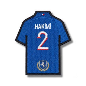 May include: A blue embroidered patch shaped like a jersey with a black border. It features the white text HAKIMI above the number 2 in white with a red outline. A small gold crest with a horse and laurel leaves is at the bottom center.