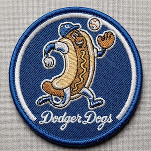 Può includere: Toppa rotonda blu con bordo bianco, raffigurante un hot dog dei cartoni animati in uniforme da baseball che lancia una palla. Il testo "Dodger Dogs" è in bianco.