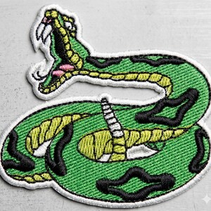 May include: Embroidered patch featuring a green snake with yellow and black markings. The snake has an open mouth with pink and white details, and is outlined in white. The patch is designed to be sewn or ironed onto fabric.