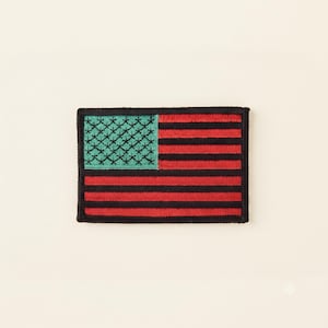 May include: A rectangular embroidered patch featuring a stylised flag design. The canton is a light teal square with black star-like symbols, while the field consists of alternating horizontal stripes in vibrant red and solid black colours.