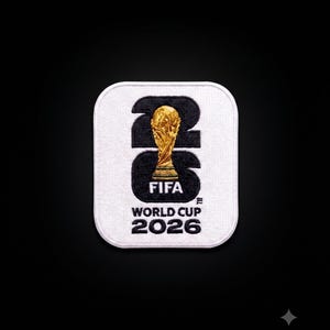 May include: A square white embroidered patch with rounded corners featuring a large black number 26. A gold-colored FIFA World Cup trophy is centered over the number. Below the trophy, the text reads FIFA WORLD CUP 2026 in black lettering.