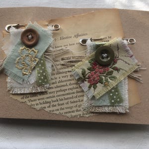 May include: Two fabric brooches attached to silver safety pins. The left brooch features light green fabric with gold embroidery and a brown button. The right brooch displays a floral pattern on cream fabric with a dark green button and green polka dot trim.