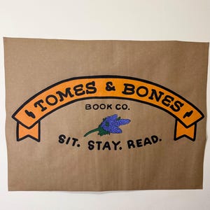 May include: A rectangular brown paper poster featuring an orange banner with the black text TOMES & BONES, the words BOOK CO. below it, a small purple flower illustration, and the black text SIT. STAY. READ. at the bottom of the design.