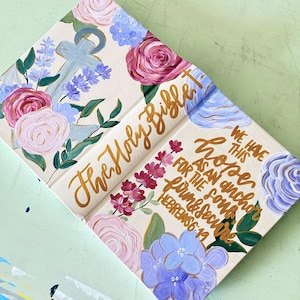 May include: A hand-painted Holy Bible with a cream background featuring pink and purple roses, blue flowers, and a light blue anchor. Gold calligraphy reads: The Holy Bible, We have this hope as an anchor for the soul, firm & secure, Hebrews 6:19.