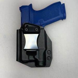 May include: A black polymer holster with a silver metal clip holds a blue handgun. The holster has a molded design with visible screw heads. The handgun is partially visible, showing its blue frame.