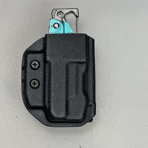 May include: A black, molded holster with two visible screws, holding a teal and silver multi-tool. The multi-tool is partially visible, showing its various components. The holster is designed to securely carry the multi-tool.