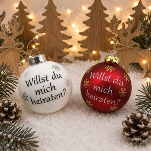 Könnte beinhalten: Zwei kugelförmige Weihnachtskugeln, eine in mattem Weiß und eine in mattem Rot, jeweils mit einer silbernen oder goldenen Kappe und dem deutschen Text "Willst du mich heiraten?" in dunkler Schrift, verziert mit kleinen Schneeflockenmotiven.