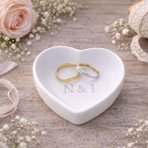 Könnte beinhalten: Eine weiße herzförmige Ringschale mit den eingravierten Initialen "N & I" hält zwei Eheringe. Ein Ring ist goldfarben, der andere silberfarben mit einem Diamanten. Die Schale ist von Blumen und Bändern umgeben.