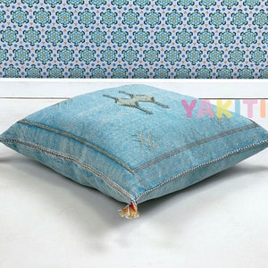 May include: A light blue square floor cushion with tassels and embroidered details. The cushion has a geometric pattern and the word "YAKITI" is visible in the background.