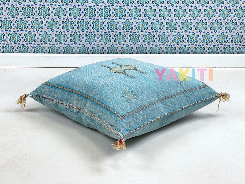 May include: A light blue square floor cushion with tassels and embroidered details. The cushion has a geometric pattern and the word "YAKITI" is visible in the background.