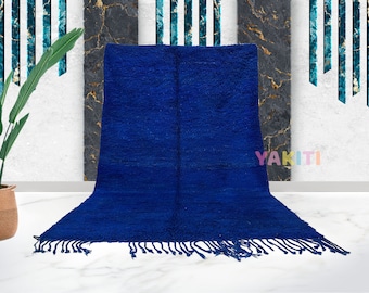 Puede incluir: Una alfombra marroquí azul oscuro con flecos en los bordes. La alfombra está colocada sobre un suelo de mármol blanco.