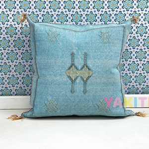 May include: A light blue throw pillow with a traditional Moroccan design. The pillow has a white background with a geometric pattern in a light blue color. The pillow is made of a soft, woven fabric.