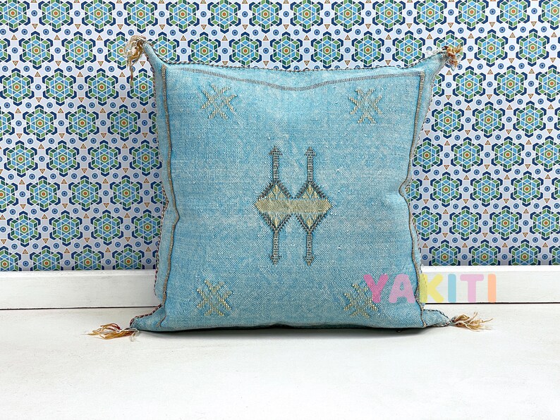 May include: A light blue throw pillow with a traditional Moroccan design. The pillow has a white background with a geometric pattern in a light blue color. The pillow is made of a soft, woven fabric.