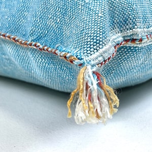 May include: A light blue denim pillow with a tassel made of white, red, and gold thread. The pillow has a decorative stitch around the edge.