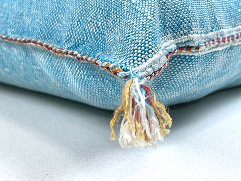 May include: A light blue denim pillow with a tassel made of white, red, and gold thread. The pillow has a decorative stitch around the edge.