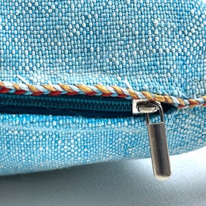 May include: A close-up of a blue pillow with a zipper closure. The zipper pull is silver and the zipper is surrounded by a decorative trim in red, white, and gold.