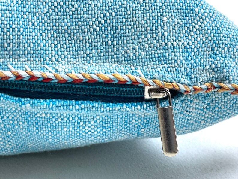 May include: A close-up of a blue pillow with a zipper closure. The zipper pull is silver and the zipper is surrounded by a decorative trim in red, white, and gold.