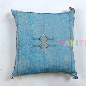 May include: A blue throw pillow with a traditional Moroccan design. The pillow has a diamond shape in the center with arrows pointing out from the sides. The diamond shape is embroidered with a white thread. The pillow has a light blue background with a subtle texture.