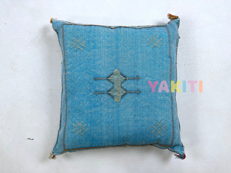 May include: A blue throw pillow with a traditional Moroccan design. The pillow has a diamond shape in the center with arrows pointing out from the sides. The diamond shape is embroidered with a white thread. The pillow has a light blue background with a subtle texture.