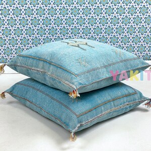 May include: Two blue denim throw pillows with tassels. The pillows are stacked on top of each other. The top pillow has a white embroidered design.