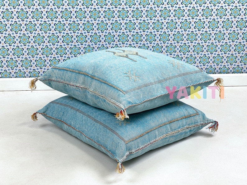 May include: Two blue denim throw pillows with tassels. The pillows are stacked on top of each other. The top pillow has a white embroidered design.