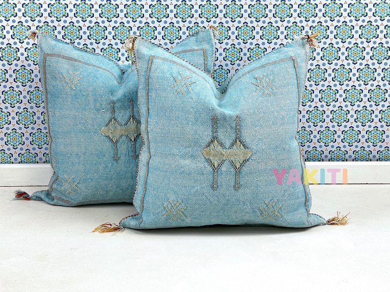 May include: Two light blue throw pillows with embroidered geometric patterns in a light beige color. The pillows have tassels on the corners.