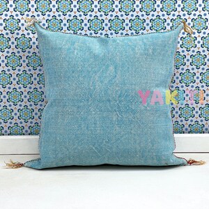 May include: A light blue square throw pillow with a subtle pattern and frayed edges. The pillow has tassels on the corners and is sitting on a white surface.