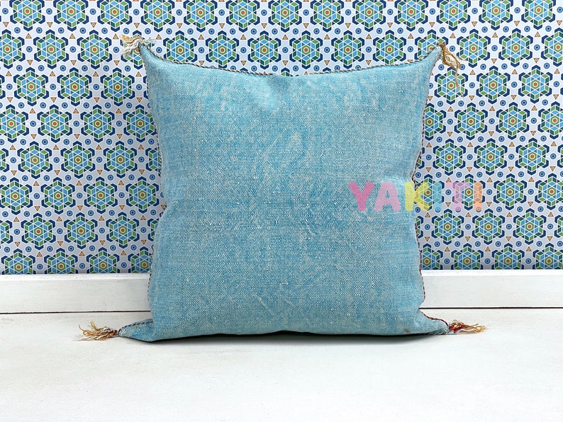 May include: A light blue square throw pillow with a subtle pattern and frayed edges. The pillow has tassels on the corners and is sitting on a white surface.