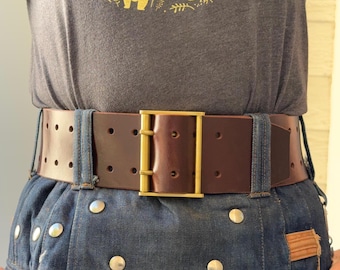 May include: A wide dark brown leather belt featuring two rows of punched holes and a rectangular brass buckle. The belt is worn over a dark gray t-shirt with a yellow bear graphic and a dark blue denim skirt with silver metal rivets.