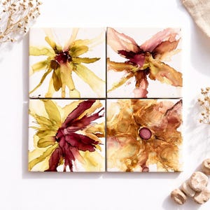 May include: A set of four square ceramic coasters, each with an abstract floral design. The coasters display watercolour-style flowers in yellow, burgundy, and brown hues on a white background. Ideal for protecting surfaces.
