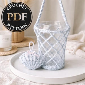 May include: A light blue crochet water bottle holder with a matching seashell-shaped coaster. The holder has a diamond pattern and a handle. The seashell coaster has a pink bow. The image includes a "CROCHET PDF PATTERN" logo.