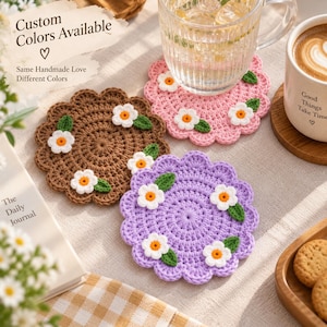 Può includere: Tre sottobicchieri rotondi all'uncinetto fatti a mano in marrone, rosa chiaro e lavanda, ciascuno con un bordo smerlato e piccoli fiori bianchi con centro giallo e foglie verdi. Il testo "Custom Colors Available" e "Same Handmade Love Different Colors" è visibile.
