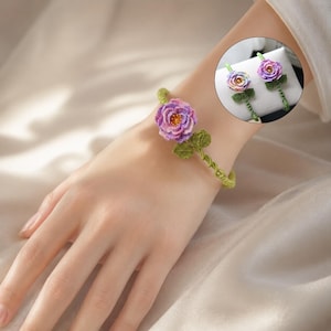 May include: A handmade bracelet and matching earrings set. The bracelet features a purple and pink flower with green leaves, attached to a green woven band. The earrings have a similar floral design. The jewelry is displayed on a wrist and a white surface.