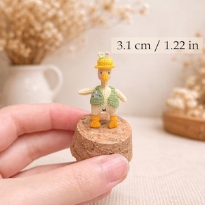 May include: A handcrafted, miniature crochet duck figurine, 1.22 inches tall, wearing a yellow hat, green vest, and orange boots. The duck stands on a cork base, showcasing intricate details and a whimsical design.